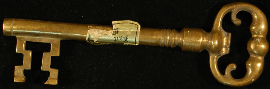 7 3/4" Bronze key with long cylindrical stem. It bears a gold foil label that reads: Fait M[ain]