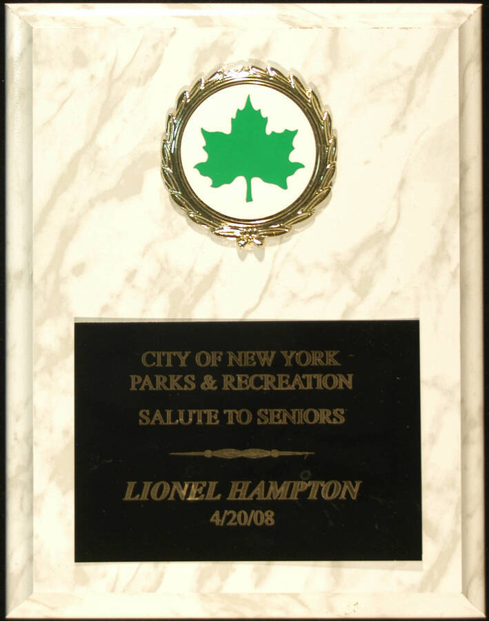 Plaque. 9"x7" White marbleized wood finish plaque with enameled white disc depicting a green leaf in a holder and engraved plate To Lionel Hampton from the City of New York Parks and Recreation. New York, NY, Apr. 20, 08[?]