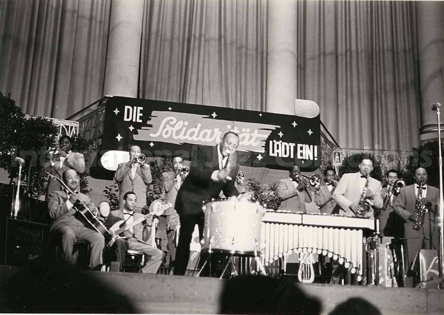 4 x 6 inch photograph. Lionel Hampton performing on the drum with orchestra, which includes guitarist Billy Mackel. A banner on the back of the stage reads: Die Solidaritat Ladt Ein!