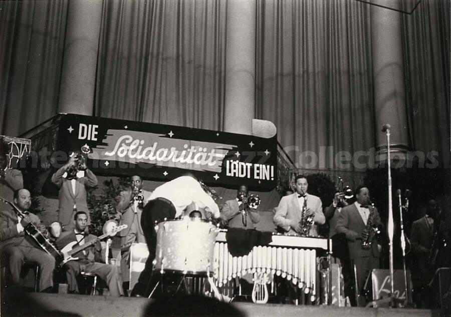 4 x 6 inch photograph. Lionel Hampton performing on the drum with orchestra, which includes guitarist Billy Mackel. A banner on the back of the stage reads: Die Solidaritat Ladt Ein!