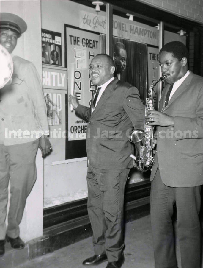 10 x 8 inch photograph. Lionel Hampton with unidentified saxophonist in front of a window which display posters announcing his performance