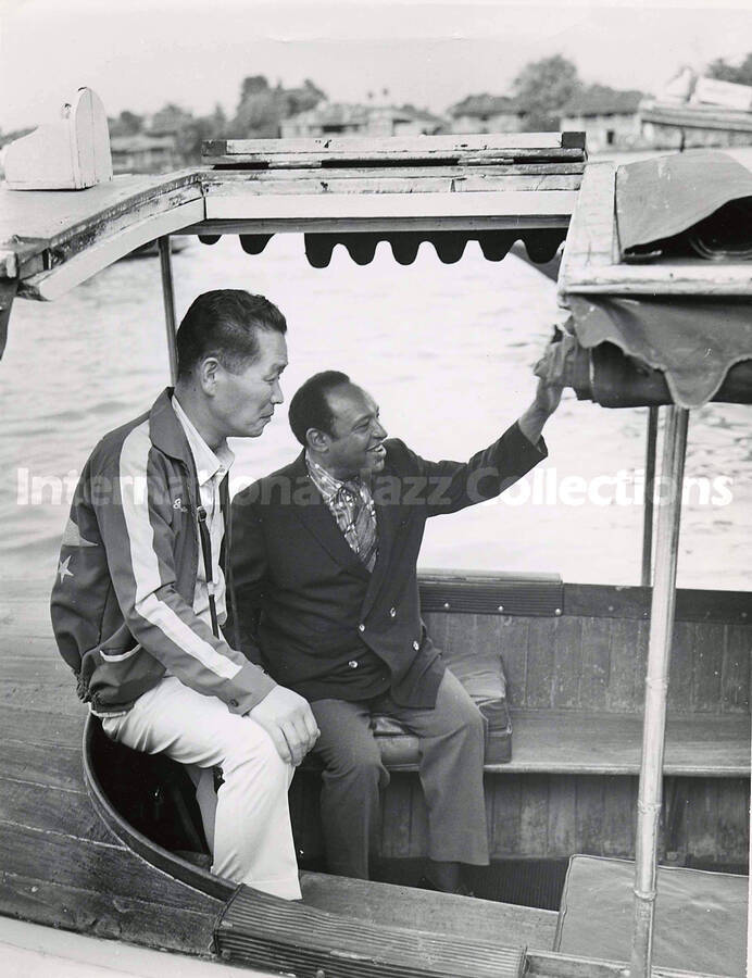 10 x 8 inch photograph. Lionel Hampton in Thailand. Typewritten note on the back of the photograph reads: Hampton cruises through The Venice of the East (Bangkok); Mr. Hampton arrived in Thailand on April 1 and was scheduled to tour the country until April 13; a photographer stands next to Mr. Hampton