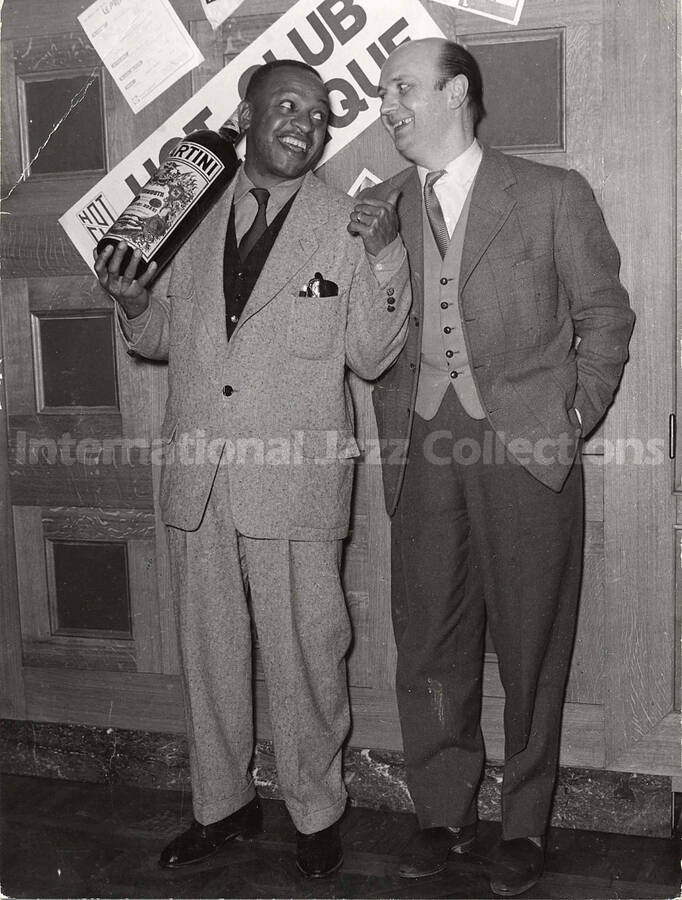 9 1/2 x 7 inch photograph. Lionel Hampton poses for Martini vermouth with unidentified man, in Brussels, Belgium. A sign on the wall reads: Hot Club Belgique