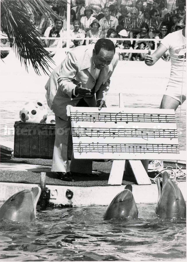 8 x 6 inch photograph. Lionel Hampton holds an enlarged musical score while standing by a pool showing it to dolphins, in front of an audience, at the Marineland. [Antibes, France?]