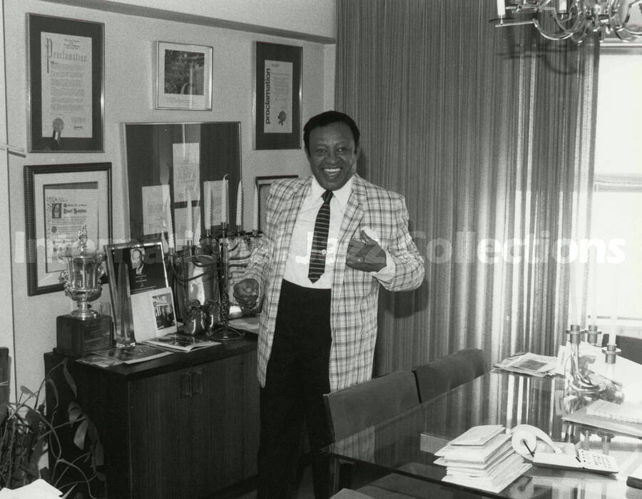8 x 10 inch photograph. Lionel Hampton poses in front of a wall displaying plaques and certificates, in his apartment