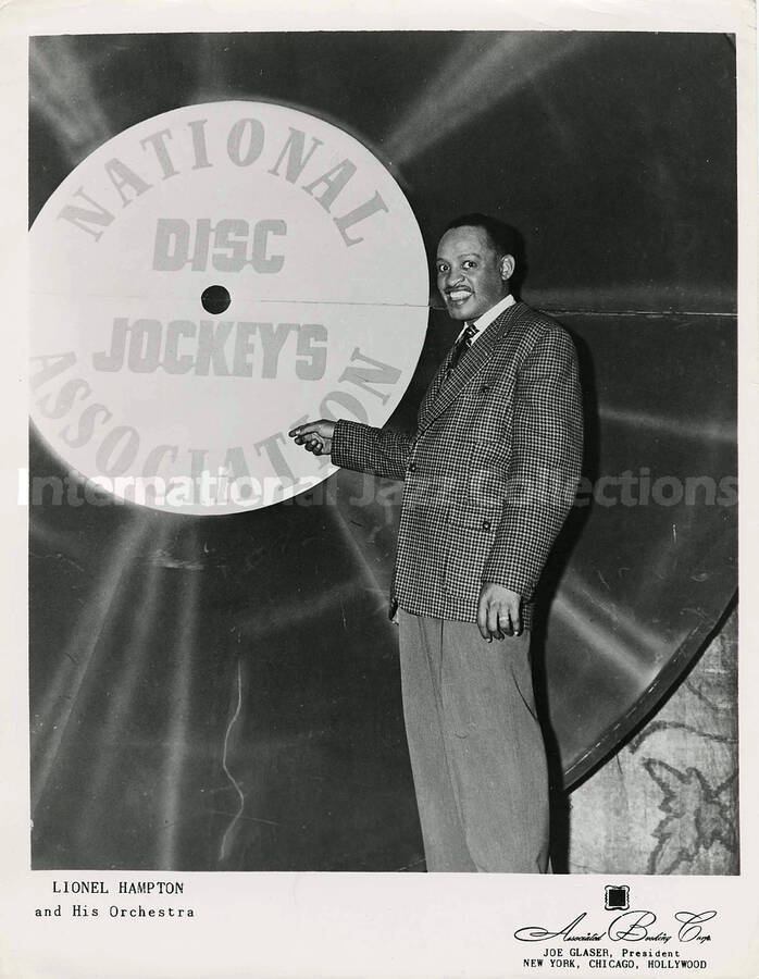 10 x 8 inch promotional photograph. Lionel Hampton poses in front of a gigantic sound record pointing to the label, which reads: National Disc Jockeys Association