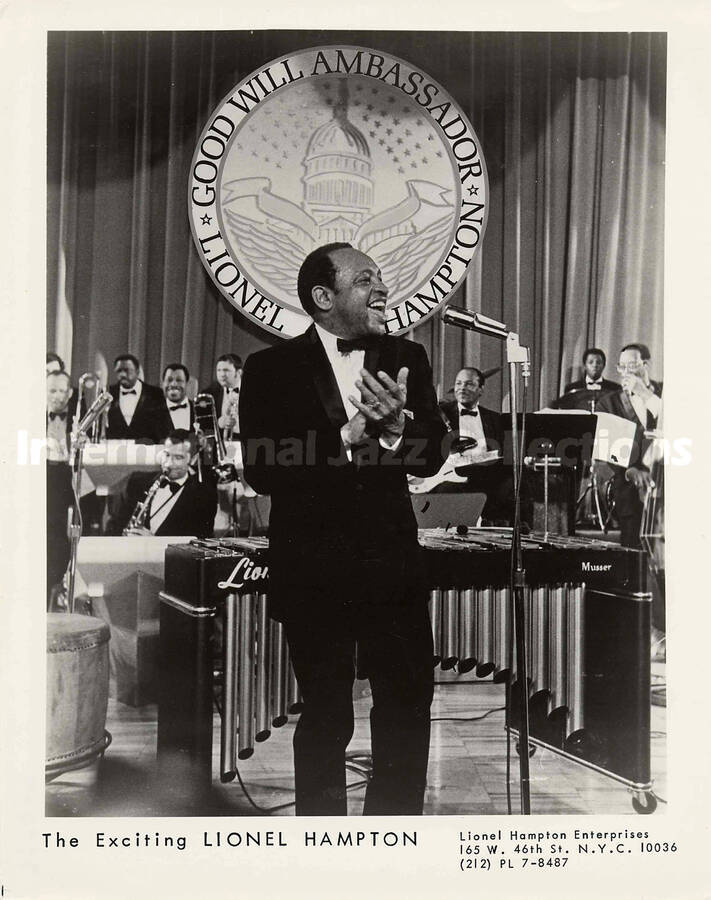 10 x 8 inch promotional photograph. Lionel Hampton standing in front of the orchestra, applauding. On the wall in the background there is a seal depicting the Capitol and the inscription: Good Will Ambassador; Lionel Hampton. Compare the foreground of this photograph with the foreground of photograph LH.III.2435