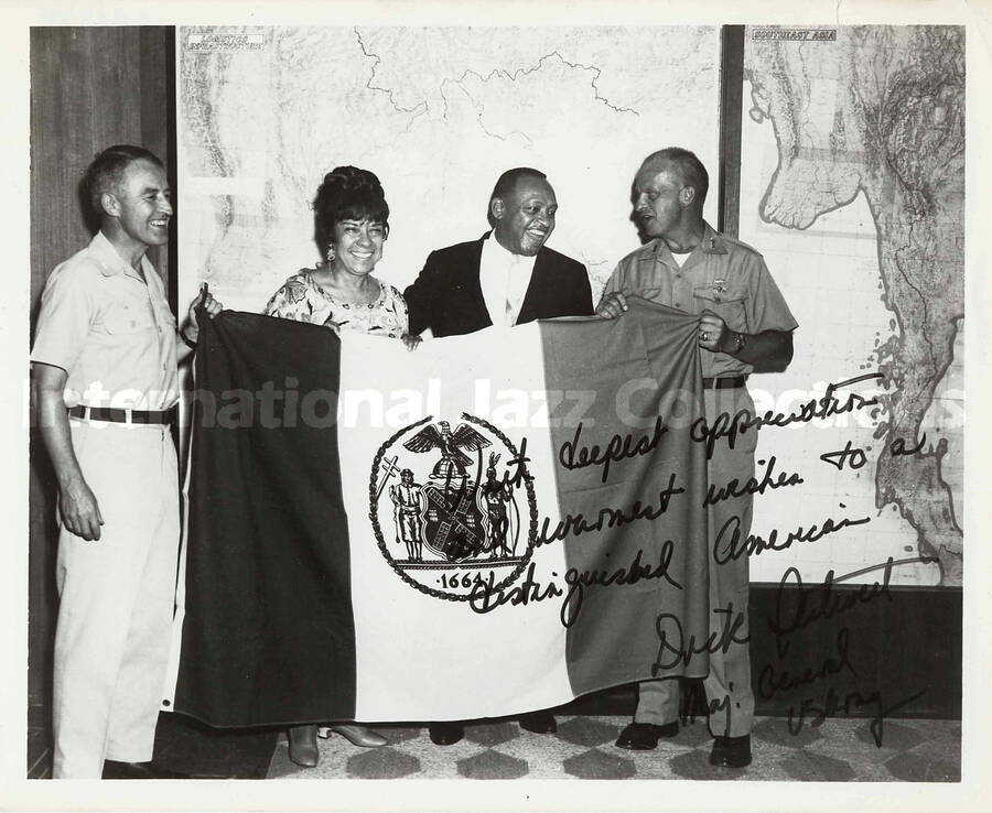8 x 10 inch photograph. Gladys and Lionel Hampton holding the New York City flag with two unidentified military men. Seen on the wall in the background is a map of the Southeast Asia