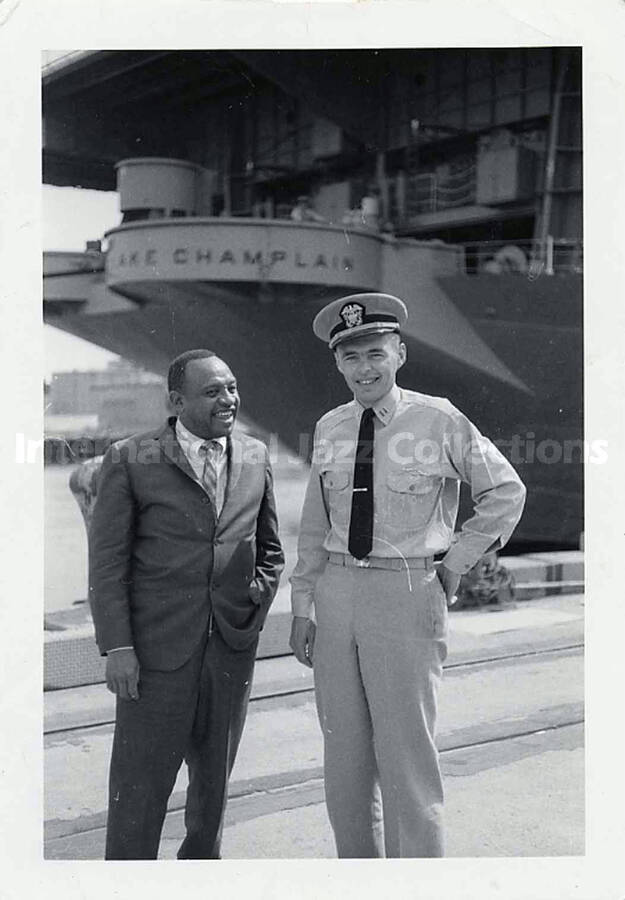 5 x 3 1/2 inch photograph. Lionel Hampton stands with and unidentified military person; The ship USS Lake Champlain CV-39 is in the background