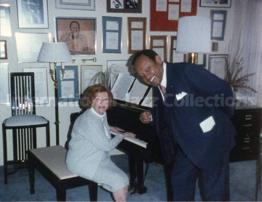 4 x 5 inch photograph. Lionel Hampton with unidentified woman sitting at the piano, in front of a wall displaying plaques and certificates, in his apartment