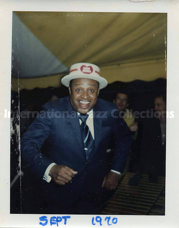 4 1/4 x 3 1/2 inch photograph. Lionel Hampton wearing a white hat with a red heart, under a tent