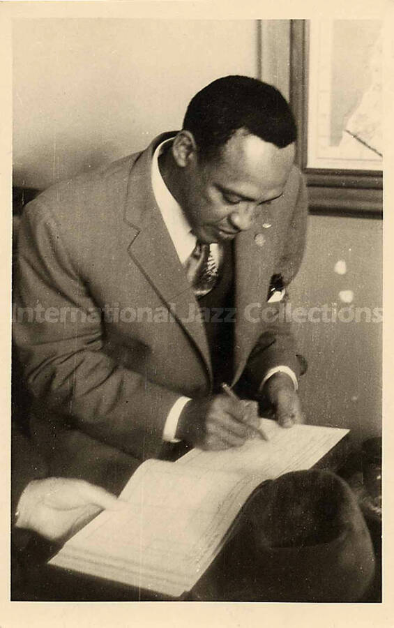 5 3/8 x 3 3/8 inch photograph in the format of postcard. Lionel Hampton writing in a calendar or ledger, [in Israel]