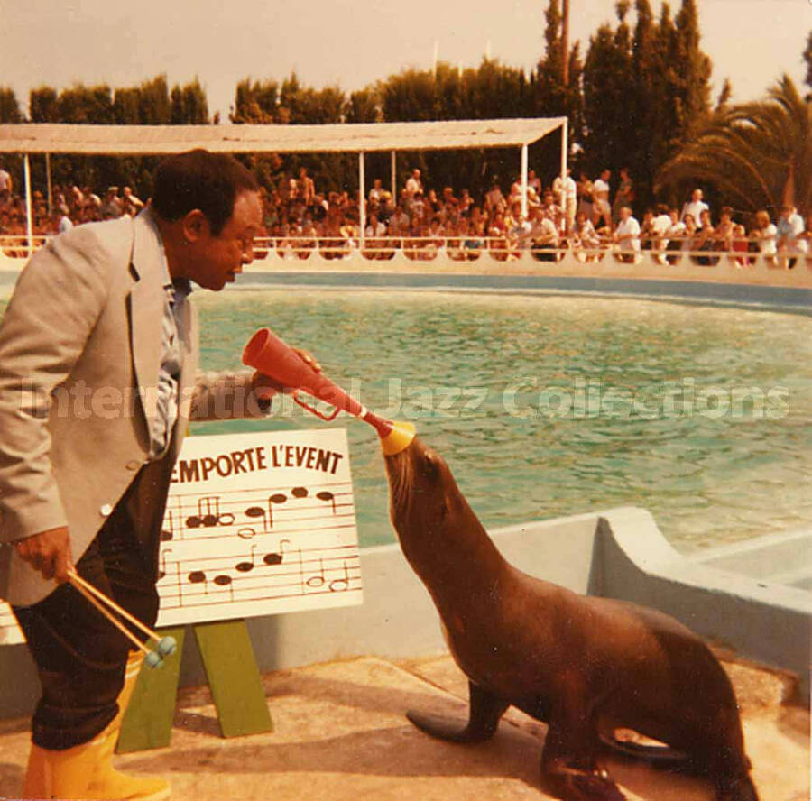 3 1/2 x 3 1/2 inch photograph. Lionel Hampton plays with a sea lion at a marine mammal park. Partially seen above a musical score are the French words: emporte l'event