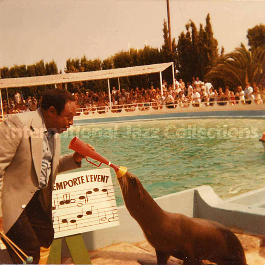 3 1/2 x 3 1/2 inch photograph. Lionel Hampton plays with a sea lion at a marine mammal park. Partially seen above a musical score are the French words: emporte l'event