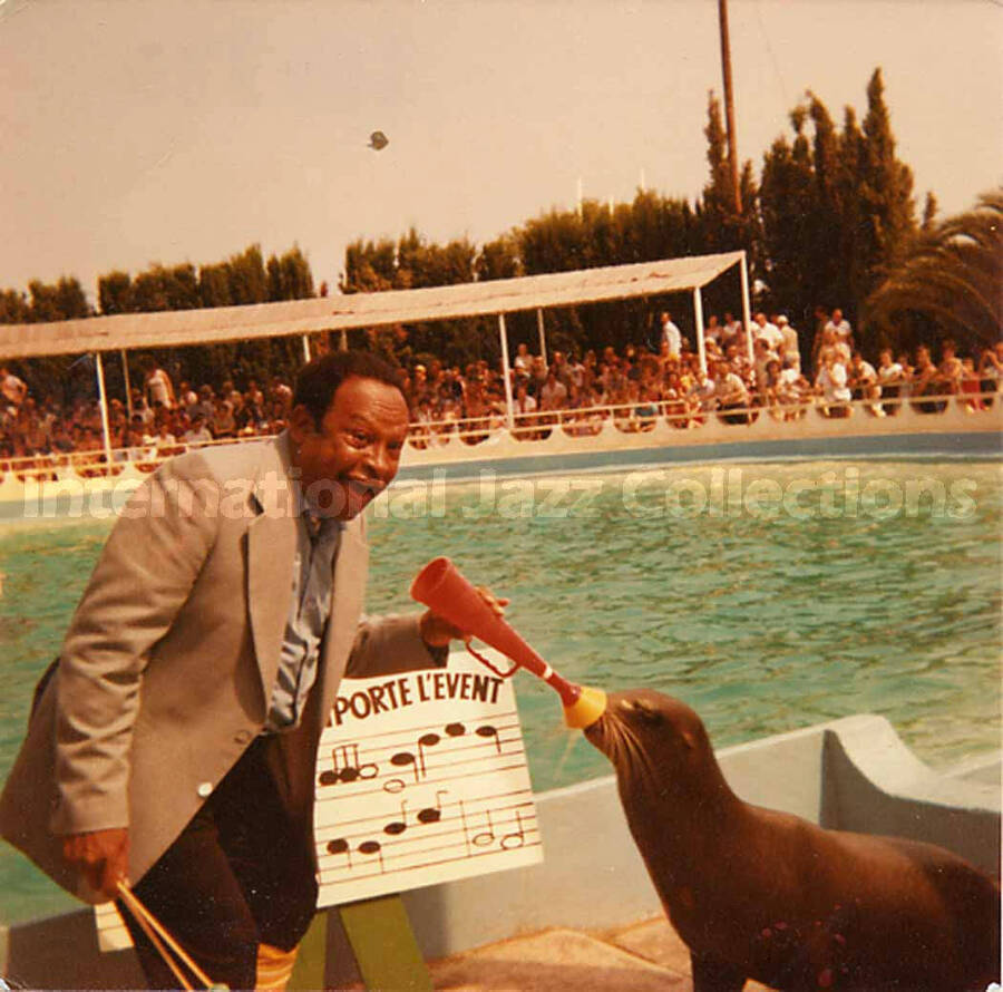 3 1/2 x 3 1/2 inch photograph. Lionel Hampton plays with a sea lion at a marine mammal park. Partially seen above a musical score are the French words: emporte l'event