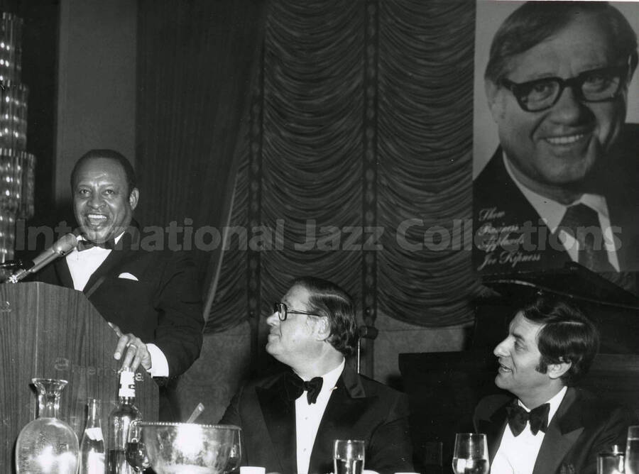 8 x 10 inch photograph. Lionel Hampton speaks observed by Joe Kipness. The podium reads: Americana. A big photo of Kipness on the wall includes the words: The business applauds Joe Kipness