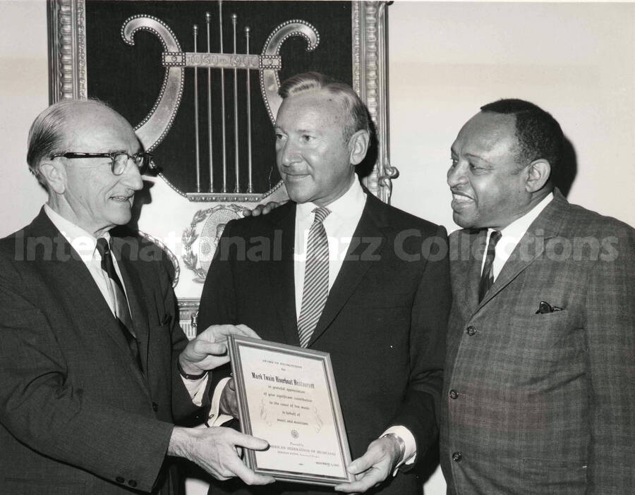 8 x 10 inch photograph. Herman Kenin, President of the American Federation of Musicians, presents an award to Jan Mitchell (center) for having booked big bands at the Mark Twain Riverboat Restaurant in N.Y., in the presence of Lionel Hampton. This photograph is accompanied by a caption note