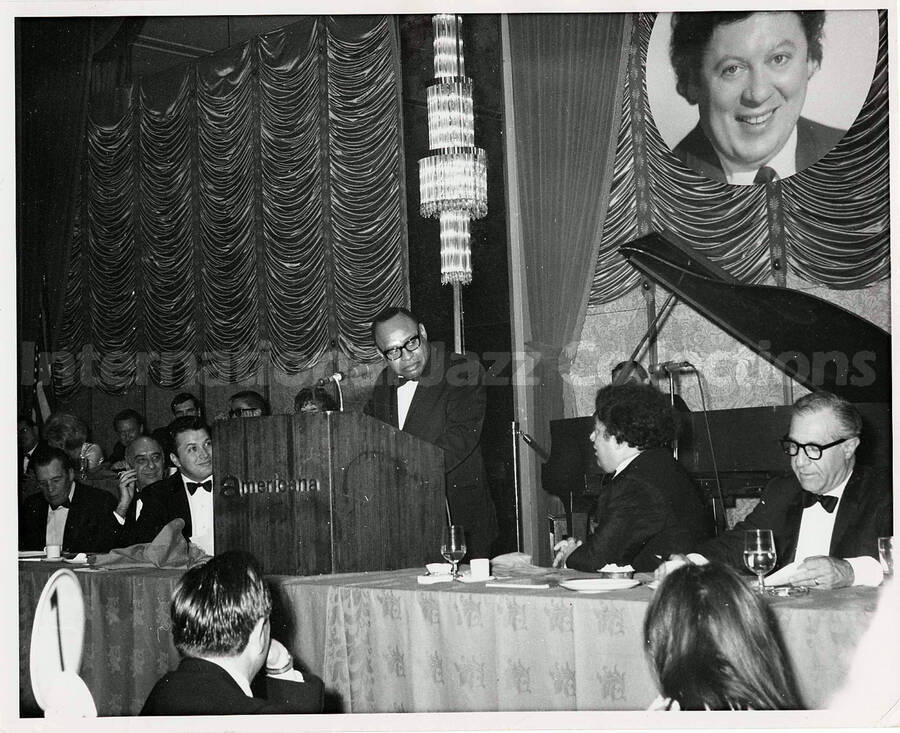 8 x 10 inch photograph. Lionel Hampton speaks observed by an unidentified man. The podium reads: Americana