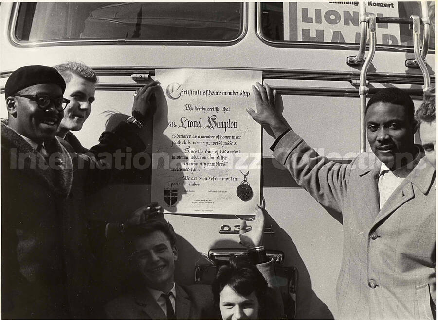 7 x 9 1/2 inch photograph. Club members of the Vienna City Ramblers Band hold certificate of Lionel Hampton's honorary membership against the back of a bus