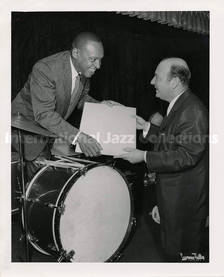 10 x 8 inch photograph. Lionel Hampton leans over drums holding a portfolio with unidentified man. There is a dedication by Don Hooker written on the drums