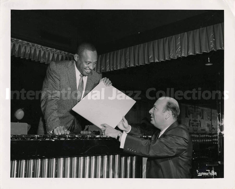 8 x 10 inch photograph. Lionel Hampton leans over vibraphone holding a portfolio with unidentified man