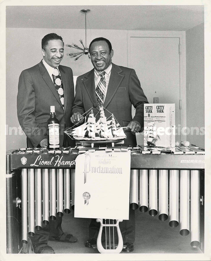 10 x 8 inch photograph. Lionel Hampton with unidentified man poses at the vibraphone with bottles of Cutty Sark whisky, in his apartment. On display in front of the vibraphone is a proclamation from the office of the Mayor of the City of New York