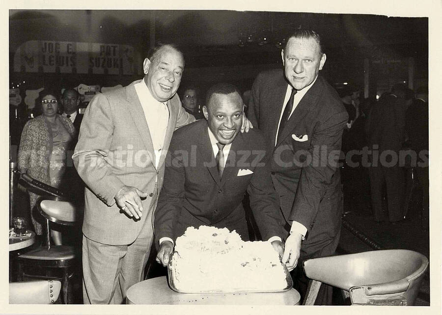 5 x 7 inch photograph. Lionel Hampton poses with a cake, sided by two unidentified men. A sign in the background reads: Joe E. Lewis; Pat Suzuki