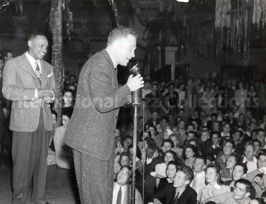 8 x 10 inch photograph. Lionel Hampton on stage observing an unidentified man at the microphone, in front an audience
