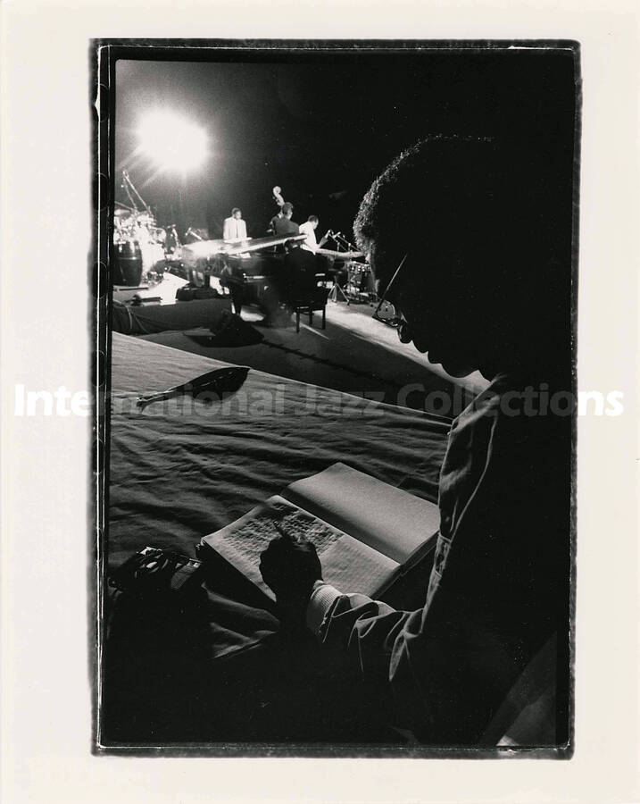 10 x 8 inch photograph. Unidentified man writes in notebook with band playing in the background