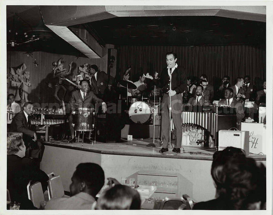 7 x 9 inch photograph. Lionel Hampton performing on drums with orchestra, which includes guitarist Billy Mackel. Joey Bishop on stage