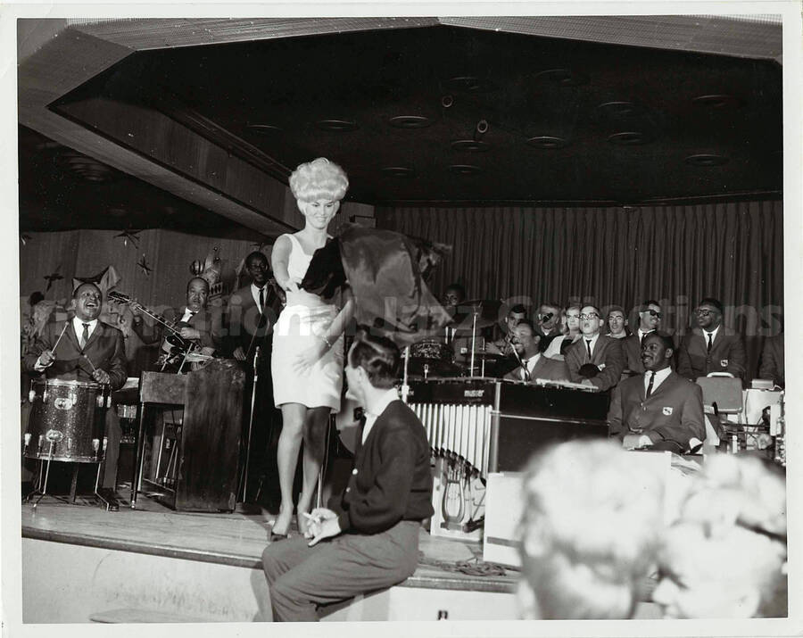 7 x 9 inch photograph. Lionel Hampton performing on drums with orchestra, which includes guitarist Billy Mackel. Joey Bishop with unidentified woman on stage