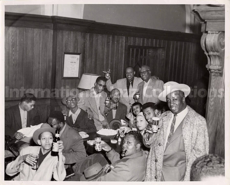 8 x 10 inch photograph. Lionel Hampton with members of his band: including singer Jimmy Scott (front left), Blues singer Sonny Parker (directly right from Scott with a pipe in his mouth), road manager George Hart (standing front right), comedian Arnold Dover (sitting directly behind Hart), Hampton (raising glass in the back right), and guitarist William Mackel (sitting on the left in front of Hampton).