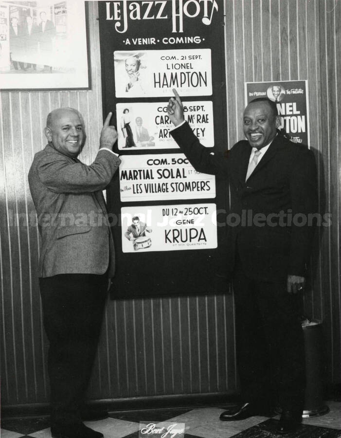 10 x 8 inch photograph. Lionel Hampton with unidentified man in front of a poster of the Le Jazz Hot. The poster, written in French, announces his performance on Sep. 21
