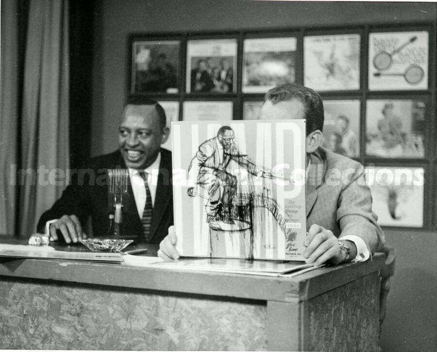 8 x 10 inch photograph. Lionel Hampton with unidentified man [in a radio studio?]. The man is holding a copy of the record The Exciting Hamp in Europe (Glad-Hamp Records)