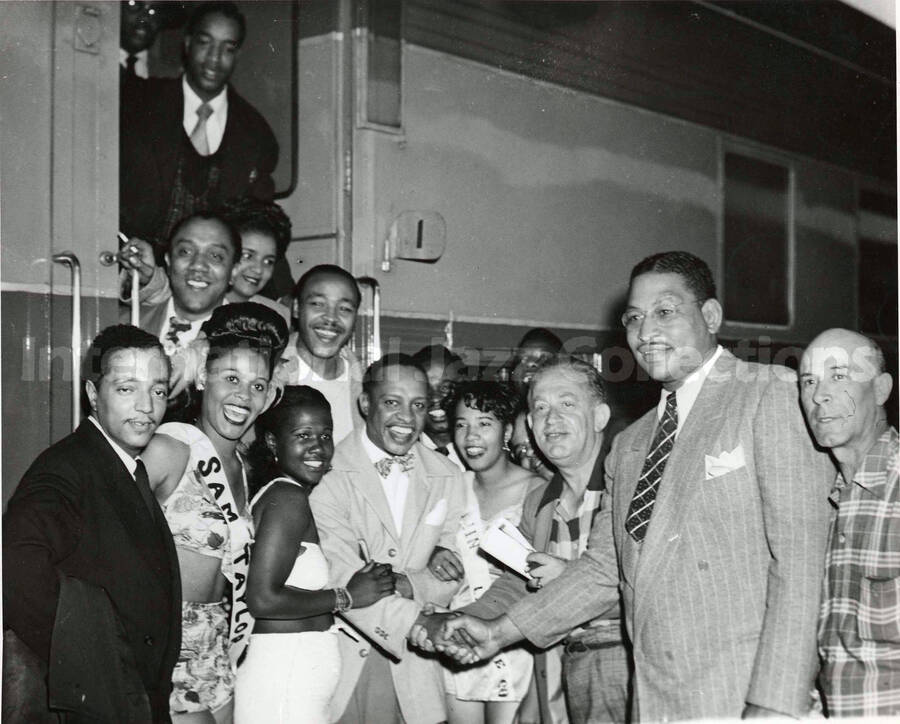 8 x 10 inch photograph. Lionel Hampton with a group of unidentified persons boarding a train, including a woman wearing a sash that reads: Sam Taylor
