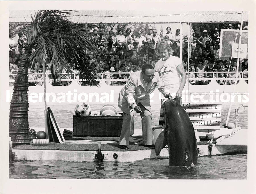 6 x 8 inch photograph. Lionel Hampton holds an enlarged musical score while standing by a pool showing it to dolphins, in front of an audience, at the Marineland. [Antibes, France?]
