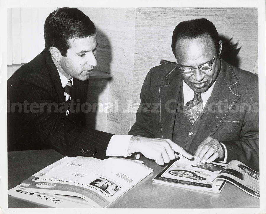 8 x 10 inch photograph. Lionel Hampton with unidentified man. The man is pointing to an Aurora skittle game on a page of a catalog. A copy of Toy & Hobby World magazine appears on the table [at the Aurora Super Fair' 74]
