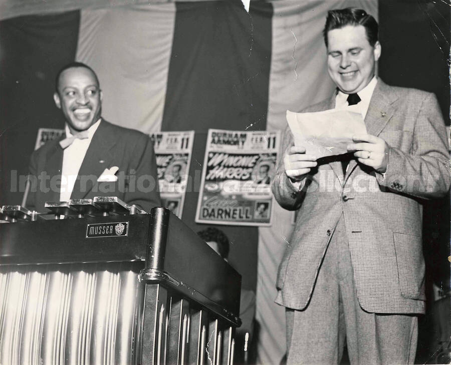 8 x 10 inch photograph. Lionel Hampton at the vibraphone with a man standing on his left reading from a piece of paper. There are three small posters in the background publicizing Wynonie Harris and [Larry] Darnell