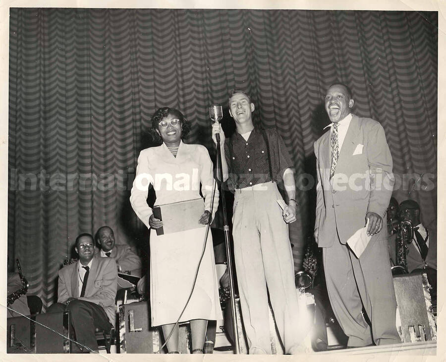 8 x 10 inch photograph. Lionel Hampton on a stage with unidentified man and woman. Musicians in the background includes guitarist Billy Mackel