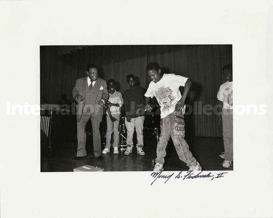 8 x 10 inch photograph. Lionel Hampton with a group of children on a stage, observing a boy dancing