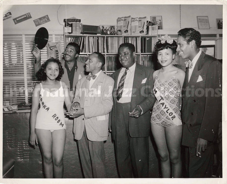 8 x 10 inch photograph. Lionel Hampton with Curley Hamner and a group of unidentified persons including two women wearing sashes. The sashes of women reads: Cobra Room and Sam Taylor