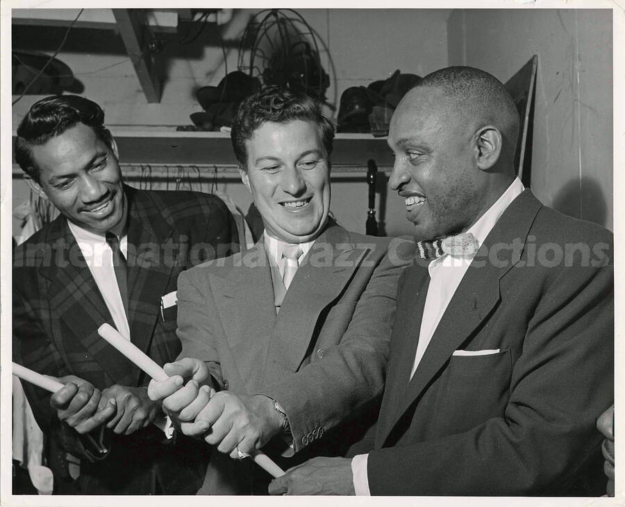 8 x 10 inch photograph. Lionel Hampton holding a drumstick in a pose of baseball player with bat alongside Curley Hamner and unidentified man