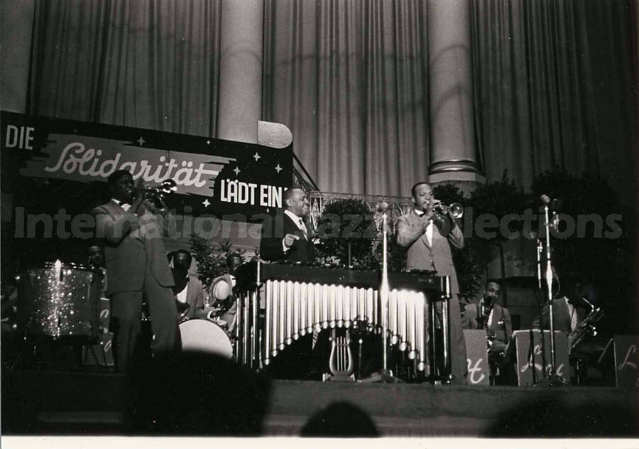 4 x 6 inch photograph. Lionel Hampton performing on the vibraphone with orchestra, which includes guitarist Billy Mackel. A banner on the back of the stage reads: Die Solidaritat Ladt Ein!