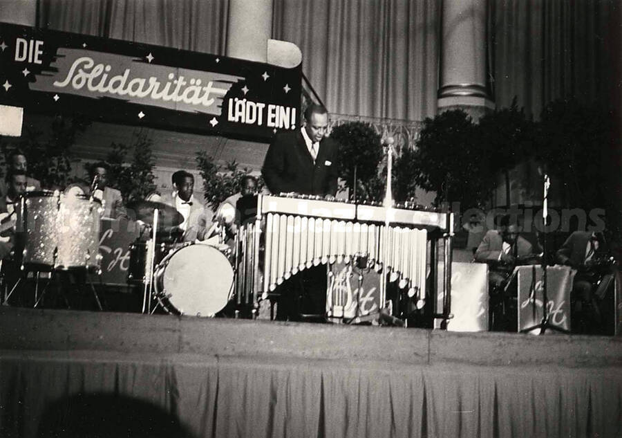 4 x 6 inch photograph. Lionel Hampton performing on the vibraphone with orchestra, which includes guitarist Billy Mackel. A banner on the back of the stage reads: Die Solidaritat Ladt Ein!