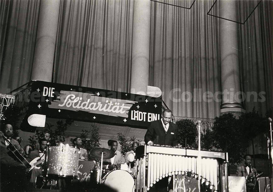 4 x 6 inch photograph. Lionel Hampton performing on the vibraphone with orchestra, which includes guitarist Billy Mackel. A banner on the back of the stage reads: Die Solidaritat Ladt Ein!