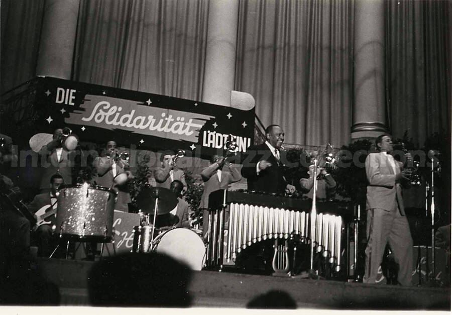 4 x 6 inch photograph. Lionel Hampton performing on the vibraphone with orchestra, which includes guitarist Billy Mackel. A banner on the back of the stage reads: Die Solidaritat Ladt Ein!