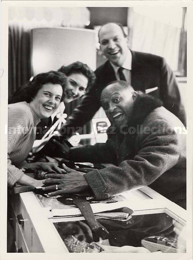 5 1/2 x 4 inch photograph. Lionel Hampton poses with unidentified persons leaning on a counter, over some ties, [in Germany?]