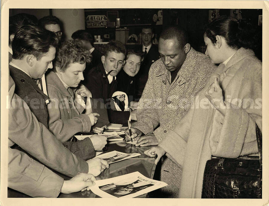 7 x 9 inch photograph. Lionel Hampton autographing for fans, in a store [abroad?]. On display in this room are photographic cameras and other articles. Seen on the background are the words: E. Leitz; Wetzlar