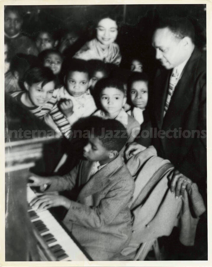 10 x 8 inch photograph. Lionel Hampton observes an unidentified boy playing the piano, surrounded by children