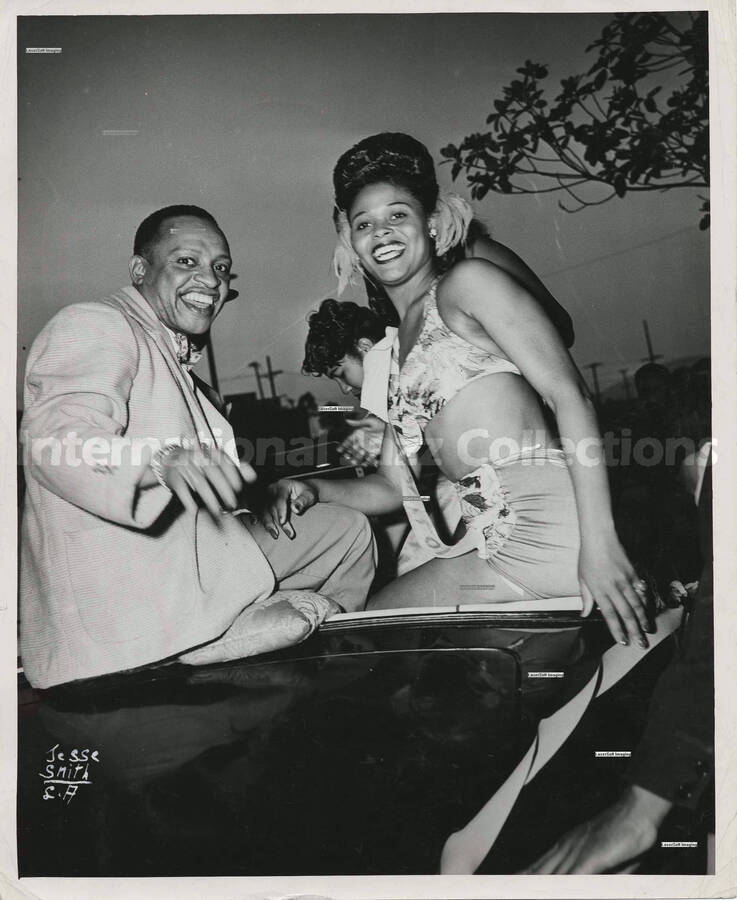 10 x 8 inch photograph. Lionel Hampton sits on the top of the bench of an open car with unidentified woman wearing a sash that reads: Sam Taylor